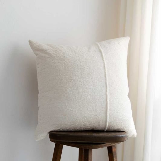 A square off-white textured throw pillow with a central vertical seam, resting on a dark wooden stool against a light wall with a curtain in the background.