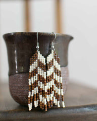 Beaded earrings with brown and white herringbone pattern on a dark surface with a blurred background