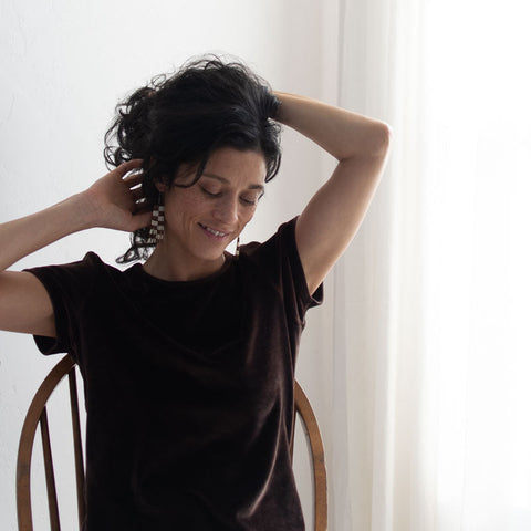 Woman sitting on a chair with her hands in her hair against a white curtain background wearing checkered beaded earrings