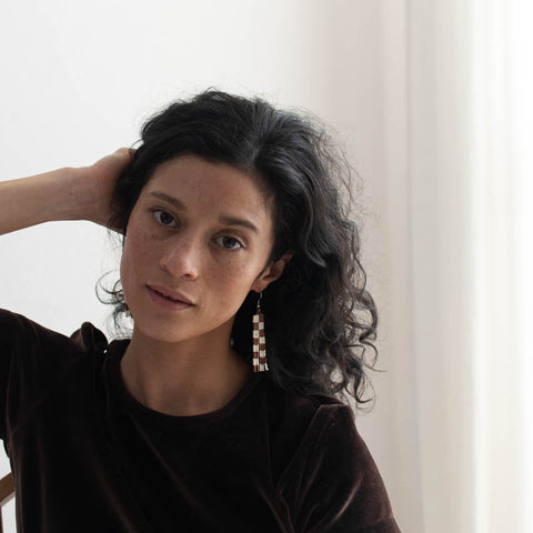 Woman with dark hair and earrings against a white curtain background modeling checkered beaded earrings