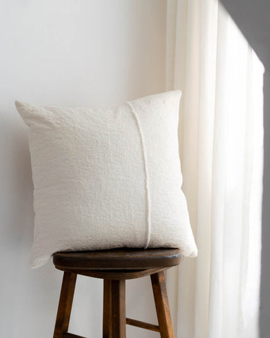 A square off-white textured throw pillow with a central vertical seam, resting on a dark brown wooden stool against a white wall next to a sheer curtain.