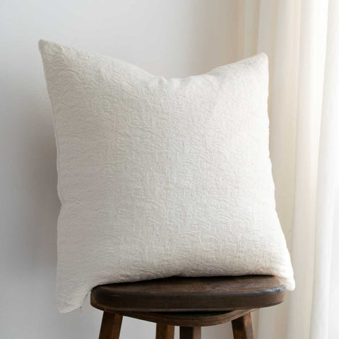 The back of a square off-white textured throw pillow resting on a dark wooden stool against a light wall with a curtain on the right.